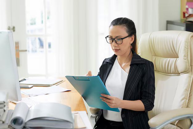 Woman in glasses, black blazer, and white top, reviews a blue clipboard at her desk in an office.