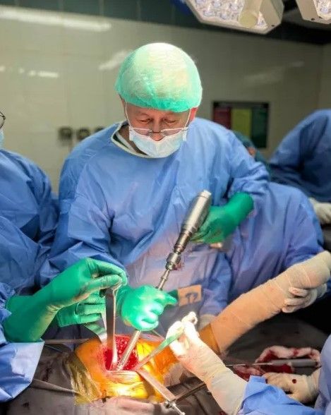 Surgeon in operating room, wearing scrubs and mask, operating on a patient's chest with medical tools.
