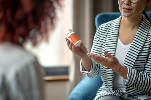 Woman in striped blazer holding medicine bottle, talking to person seated in front of her.