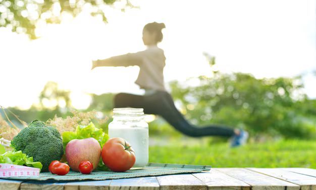 Vegetables, fruit, and yogurt on a wooden table with a blurred woman stretching in the background outdoors.