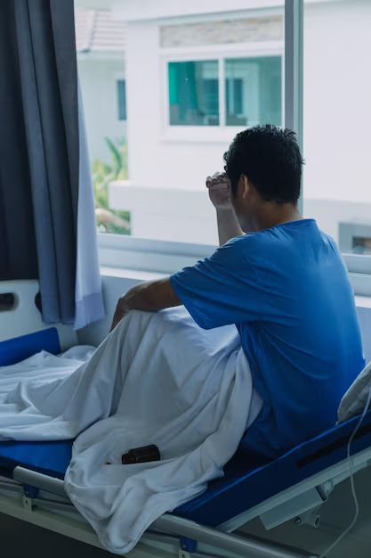 Man in blue hospital gown sits on bed, looking out window, hand on forehead.