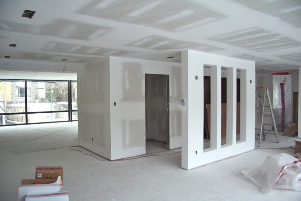 Habitación interior blanca sin terminar con paredes de pladur, grandes ventanales y una partición central en construcción.