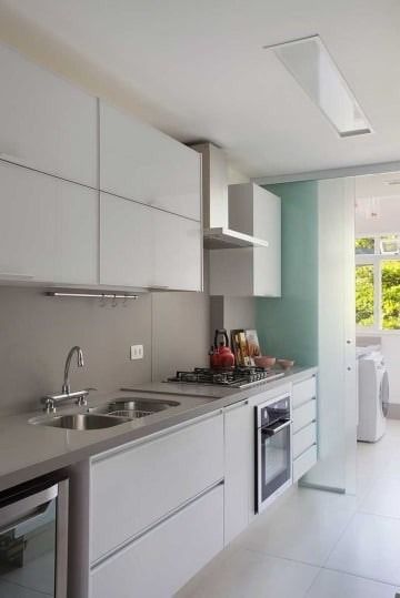 Cocina moderna blanca con encimeras grises, horno empotrado, fregadero y luz natural que entra por la ventana.