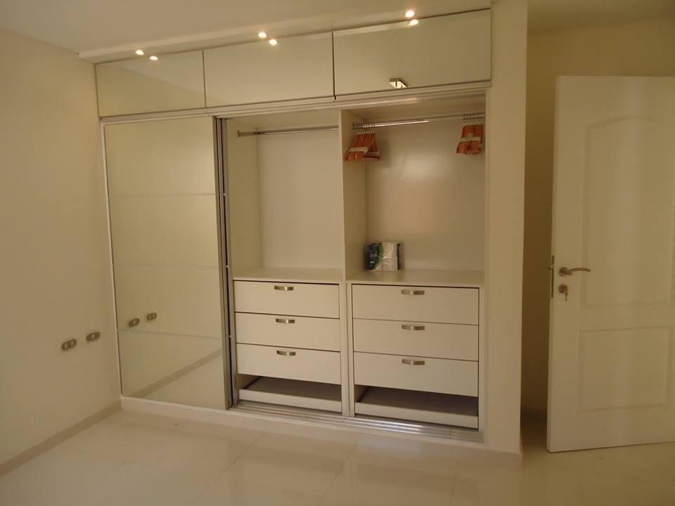 Armario empotrado blanco con puertas de espejo en un dormitorio beige, una puerta abierta que deja ver los cajones y estantes.