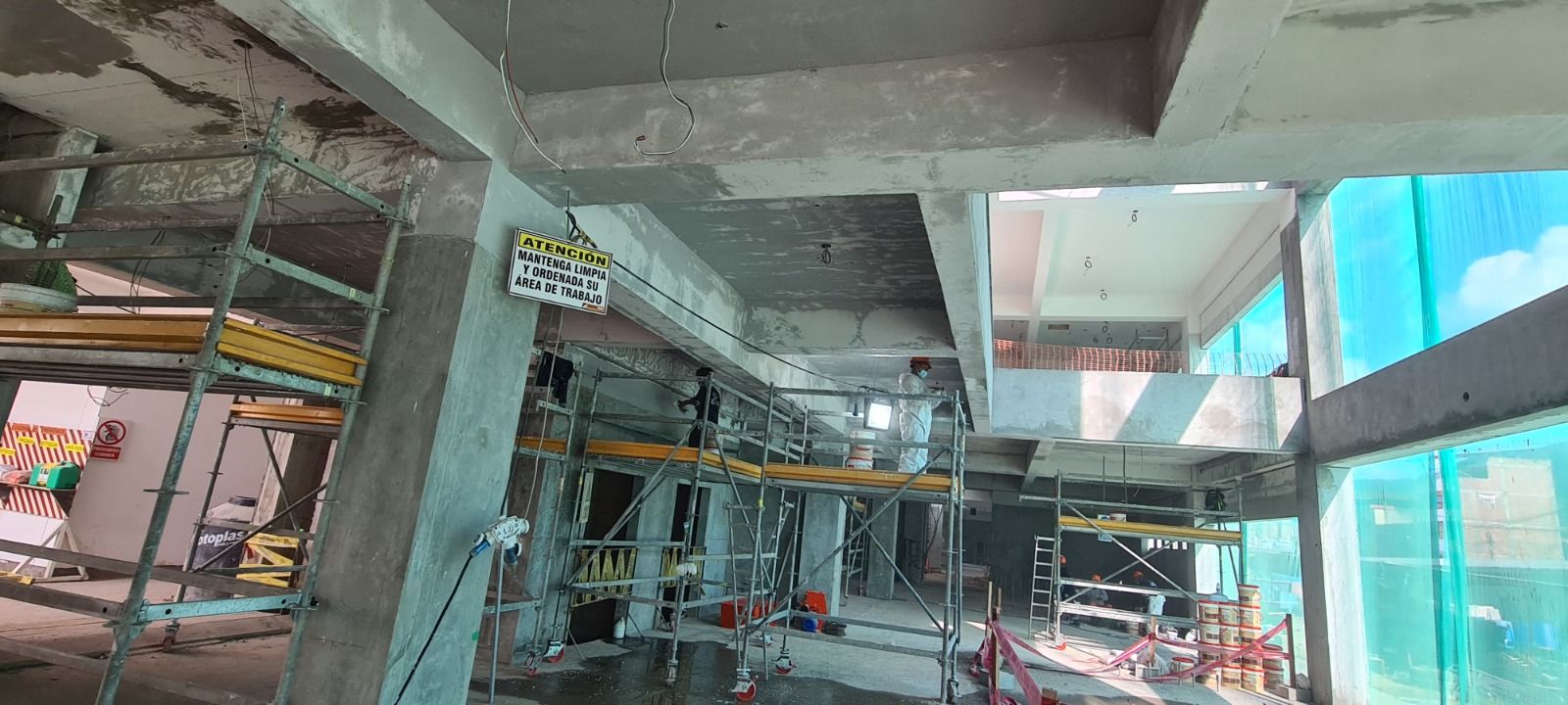 Interior de una obra en construcción con vigas de hormigón, andamios y lonas azules a lo largo de un pasillo.