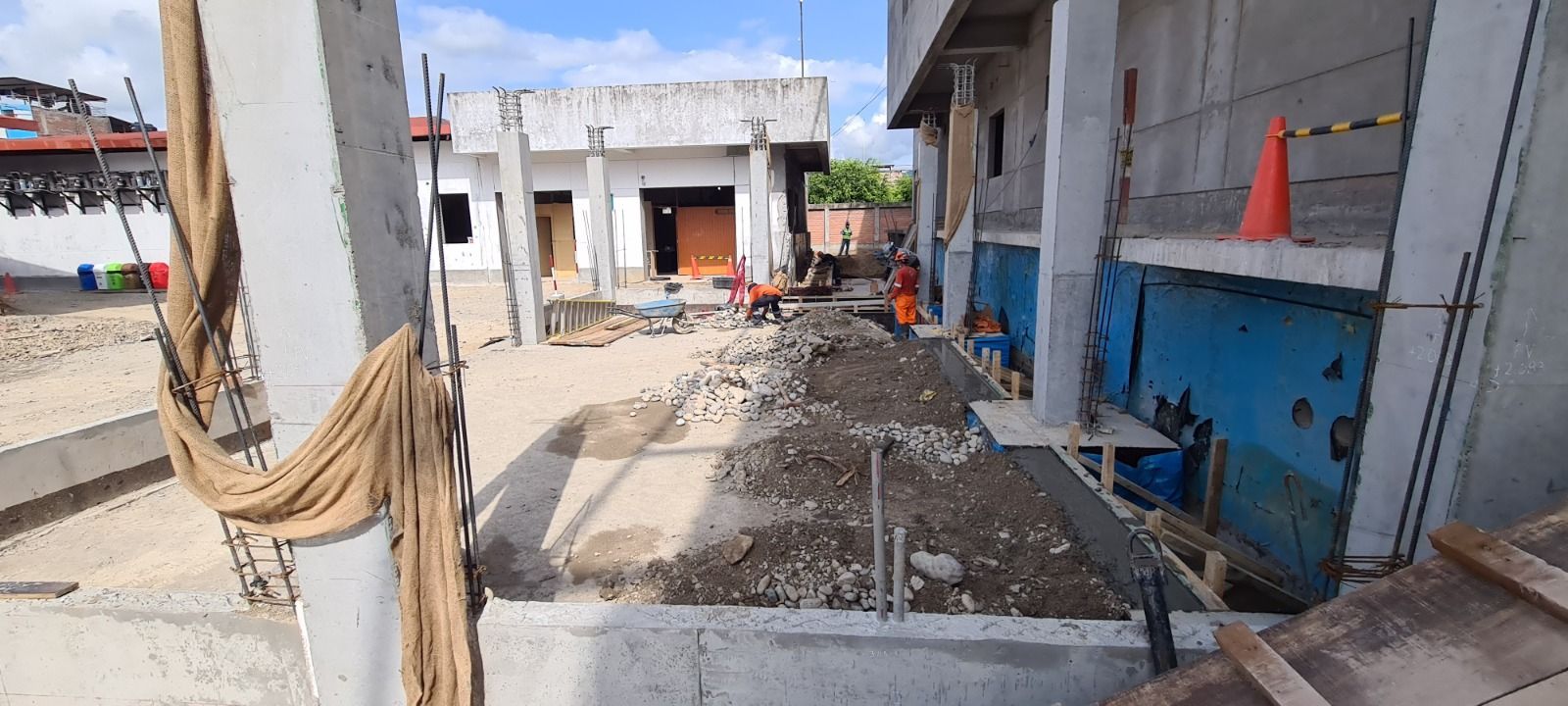 Obra en construcción con muros de hormigón, zanjas y conos de seguridad naranjas junto a un edificio.