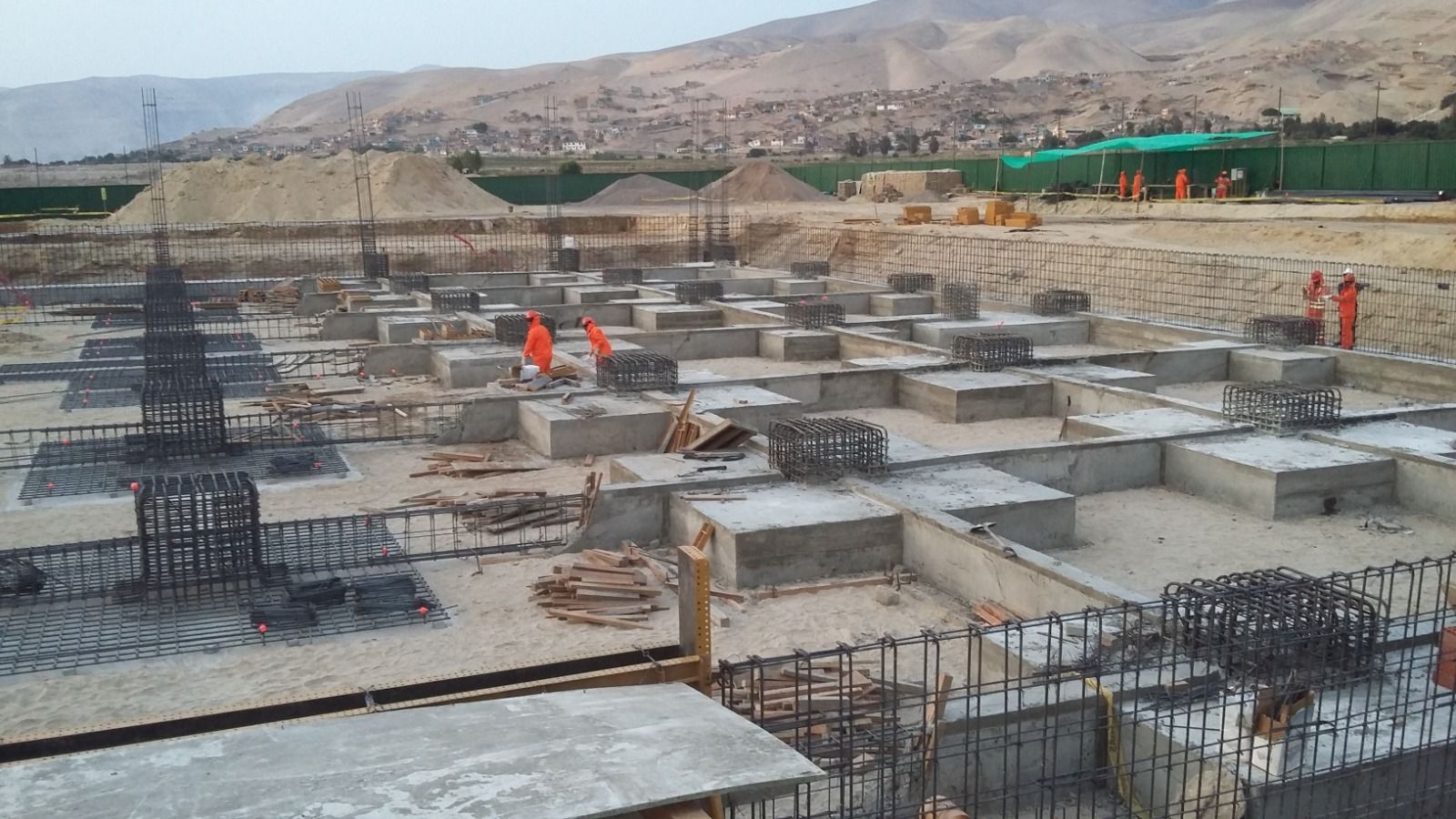 Obra en construcción con cimientos de hormigón y barras de refuerzo a la vista, trabajadores con chalecos naranjas, montañas al fondo.
