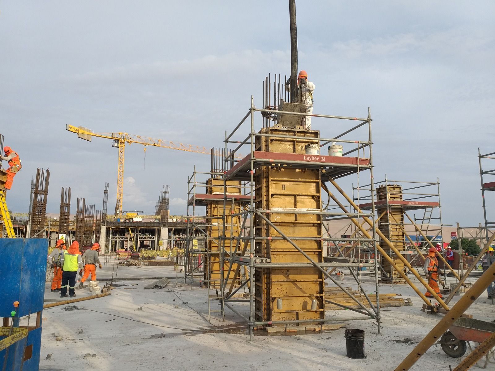 Obra en construcción con trabajadores, andamios, grúas y una columna central de hormigón en construcción.