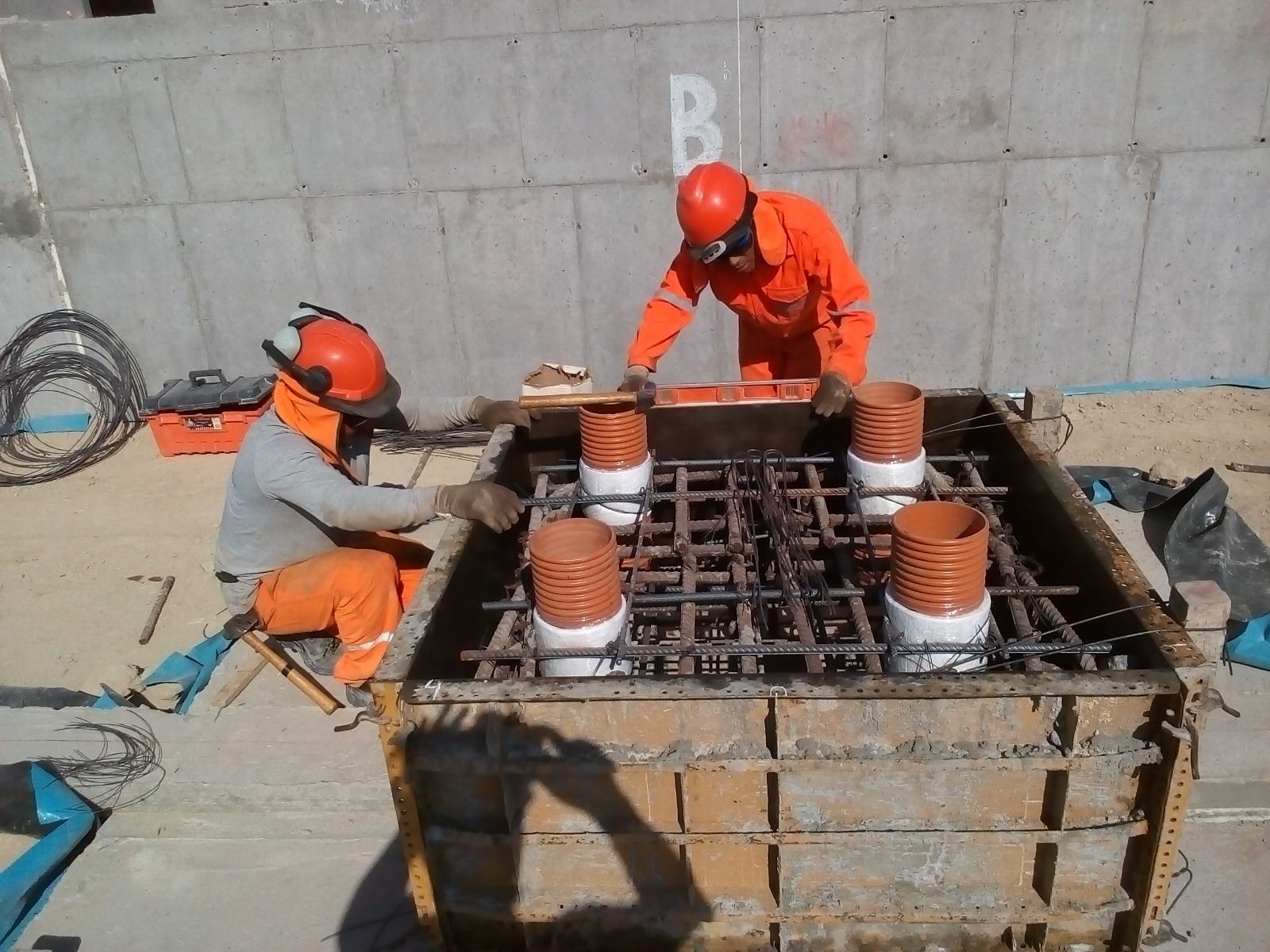 Dos trabajadores con trajes de seguridad naranjas ensamblan barras de refuerzo dentro de un encofrado de cimentación de hormigón.