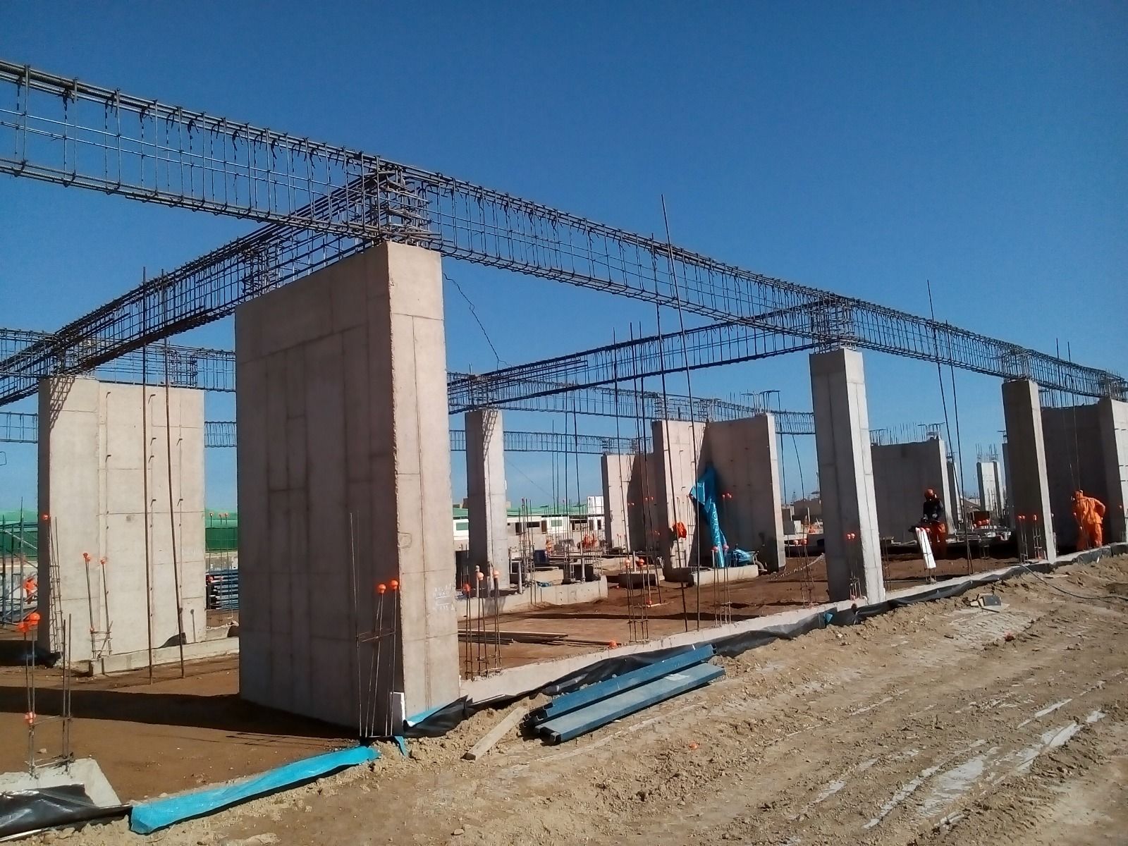 Obra en construcción con pilares de hormigón y cerchas de acero bajo un cielo azul despejado.