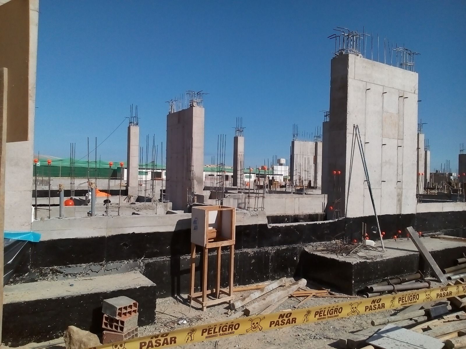 Obra en construcción con pilares de hormigón y barras de refuerzo que se elevan desde una base parcialmente construida bajo un cielo azul despejado.
