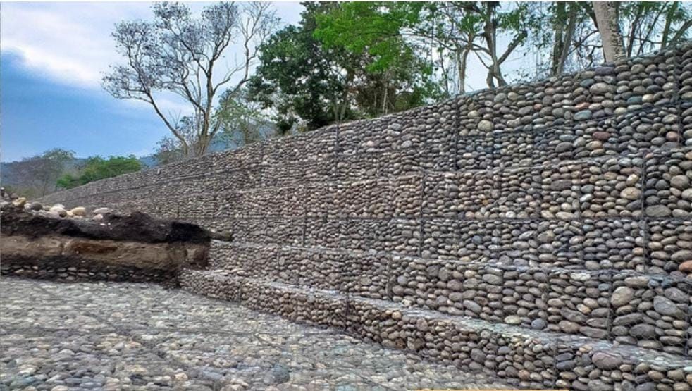 Muro de contención de piedra a lo largo de un camino empedrado, con árboles y cielo azul al fondo.