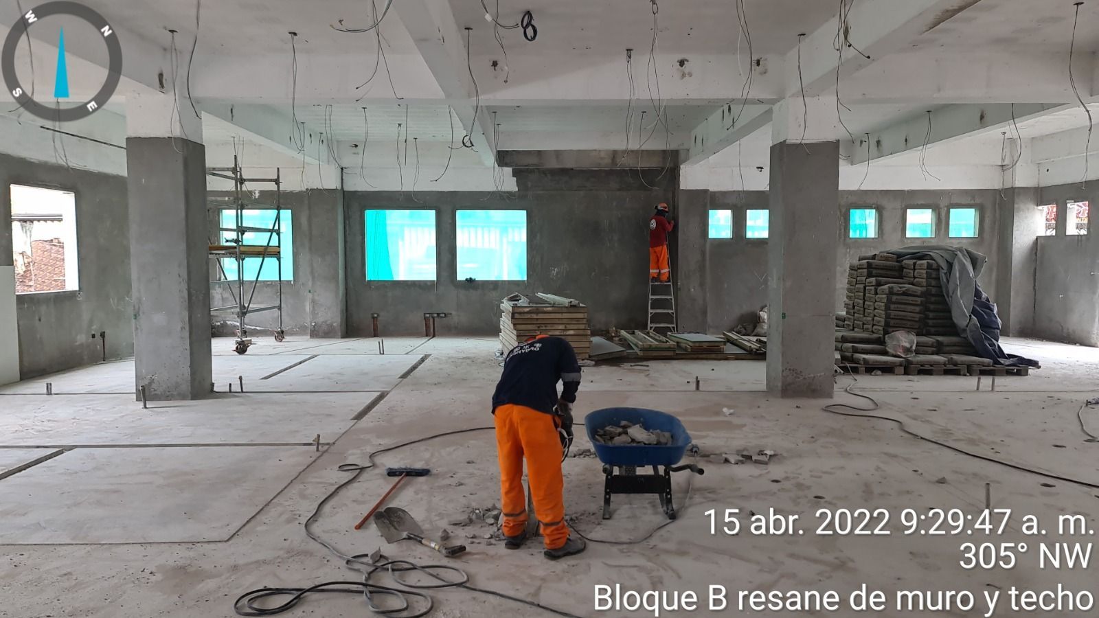 Obrero con pantalones naranjas lijando un piso de concreto en un edificio sin terminar con paredes y ventanas expuestas.