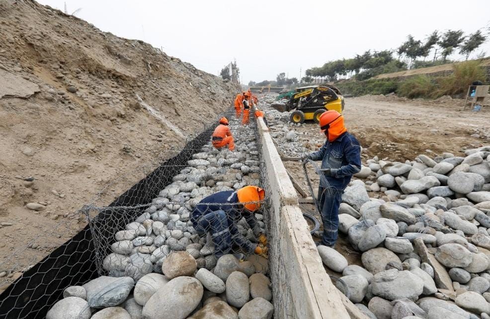 Obreros con cascos naranjas construyen un canal revestido de piedra junto a una pendiente de tierra.