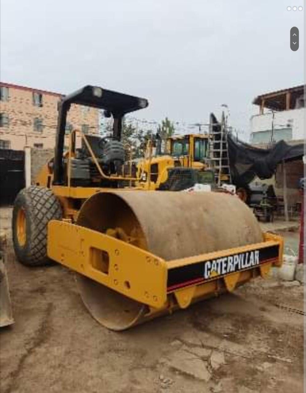 Una apisonadora Caterpillar amarilla estacionada en un patio de construcción con otra maquinaria pesada cerca.