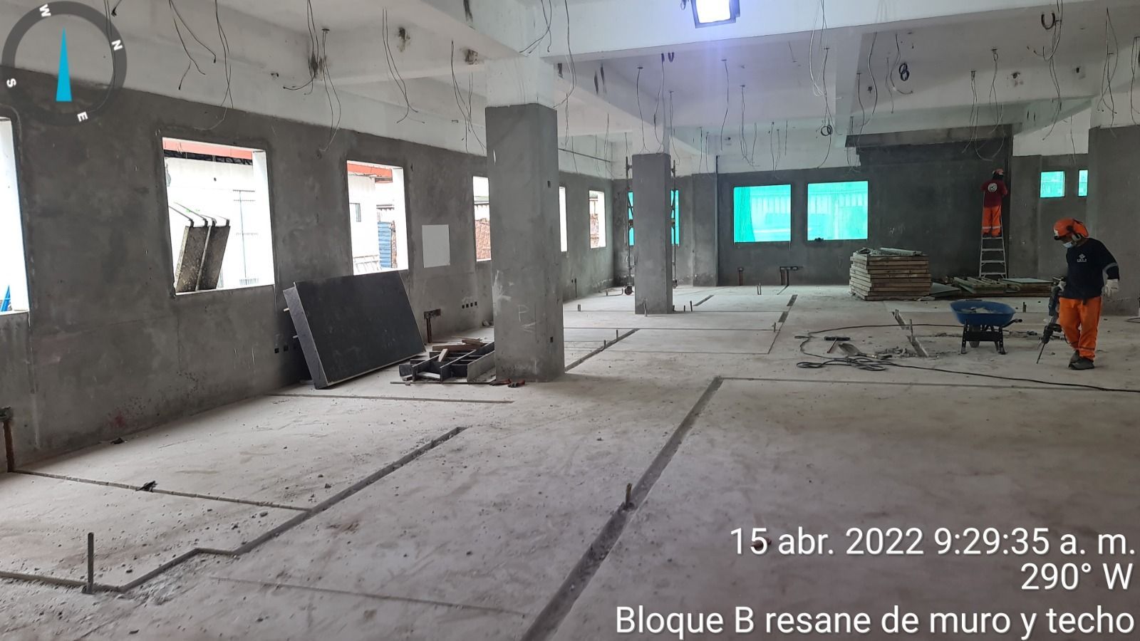 Habitación vacía de hormigón en construcción, con columnas, ventanas, materiales dispersos y un trabajador vestido de naranja a la derecha.