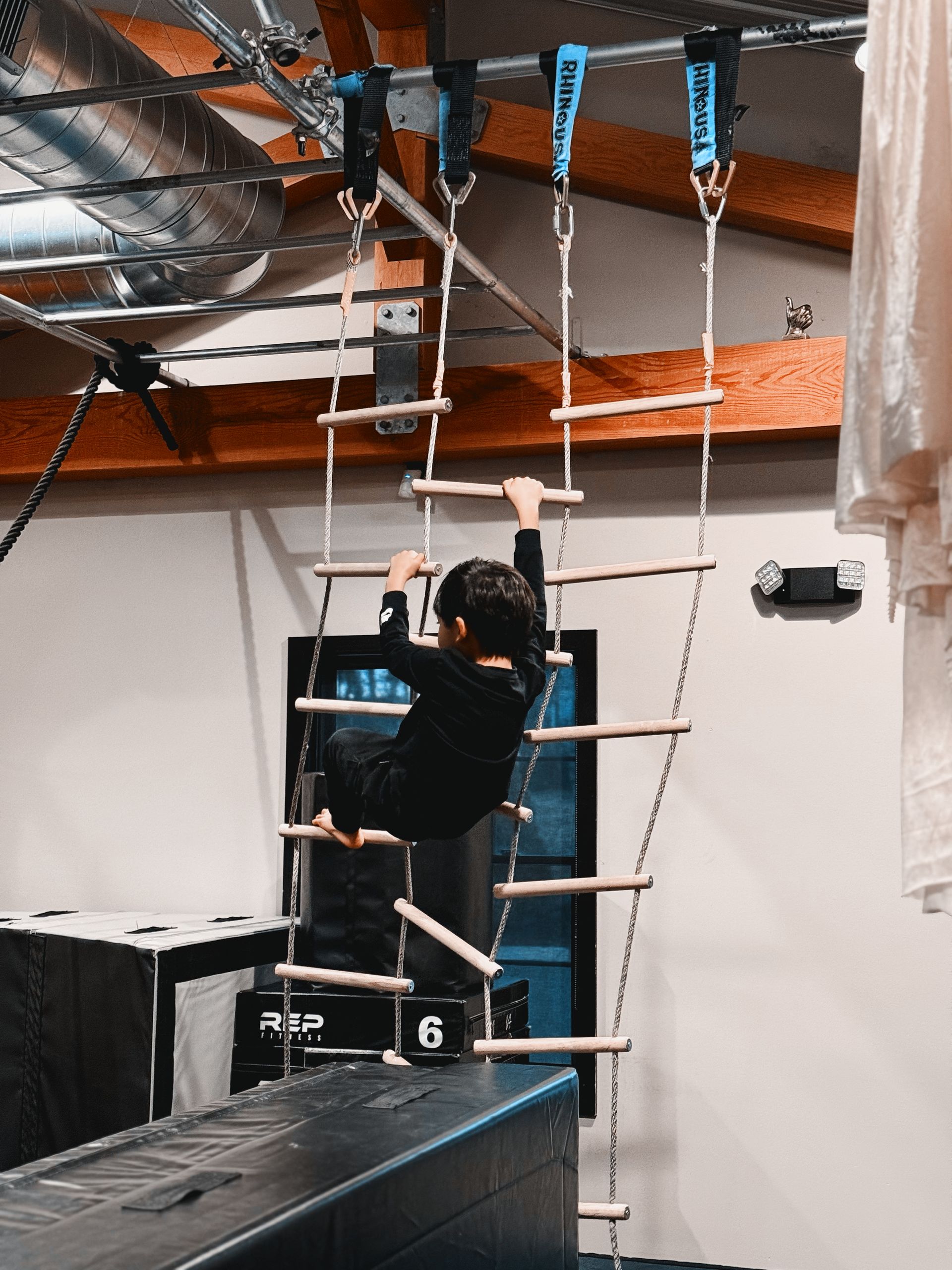 Person climbing a rope ladder obstacle course indoors, reaching upward on a black platform.