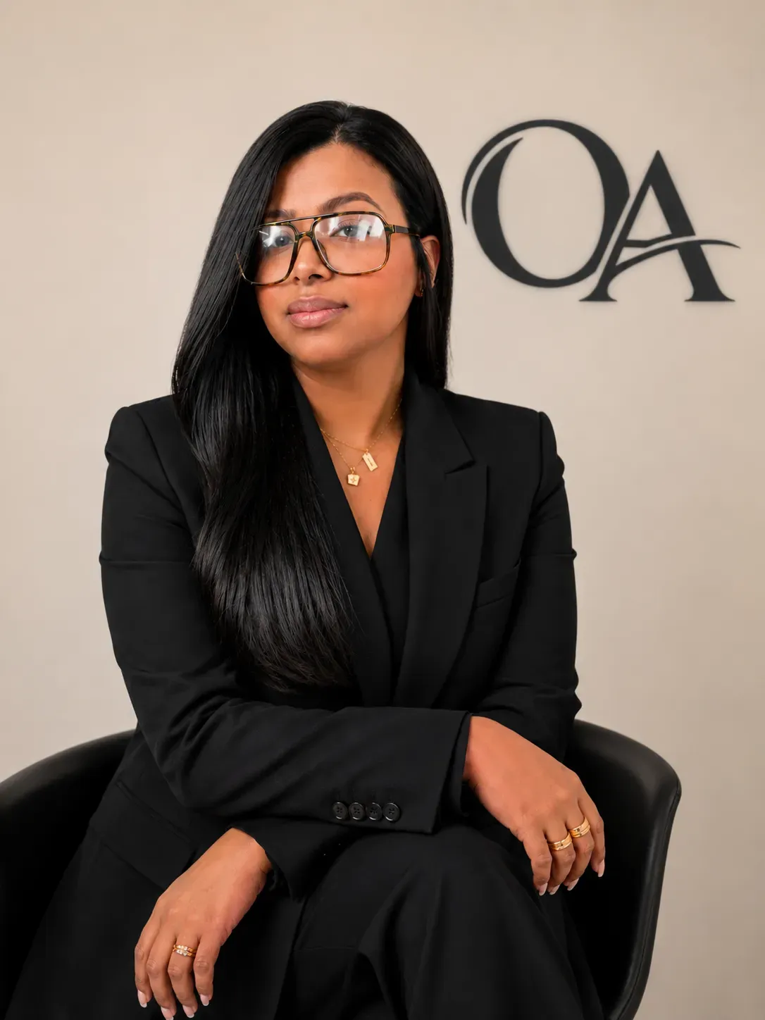 Woman in black suit and glasses seated in a chair against a beige wall with “OA” lettering.