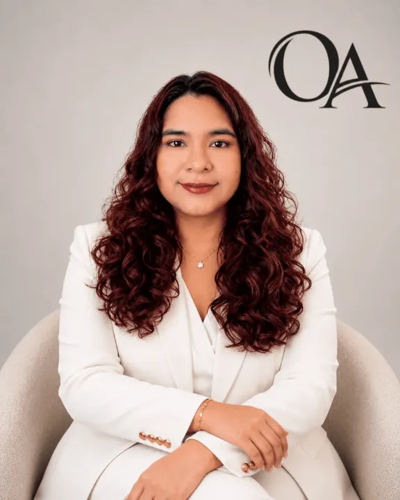 Portrait of a woman in a white blazer seated against a light background with an “OA” logo