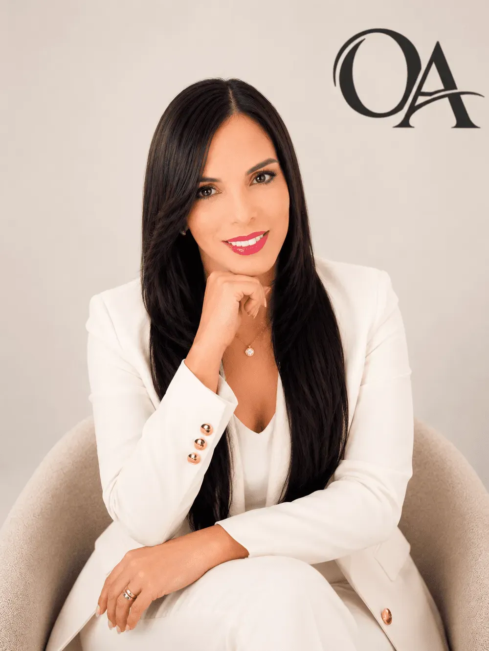 Portrait of a woman in a white blazer seated against a beige background with “OA” logo