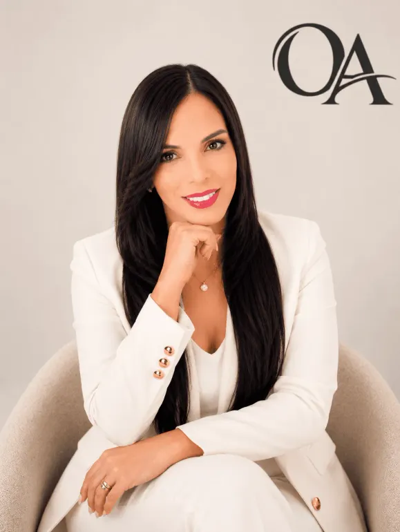 Portrait of a woman in a white blazer seated against a beige background with “OA” logo