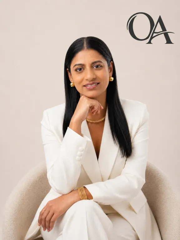 Portrait of a woman in a white suit seated against a beige background with “OA” in the corner