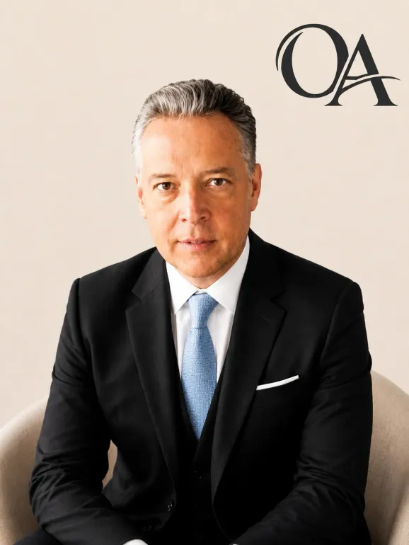 Studio portrait of a man in a black suit and blue tie, seated against a beige background with “QA” in the corner