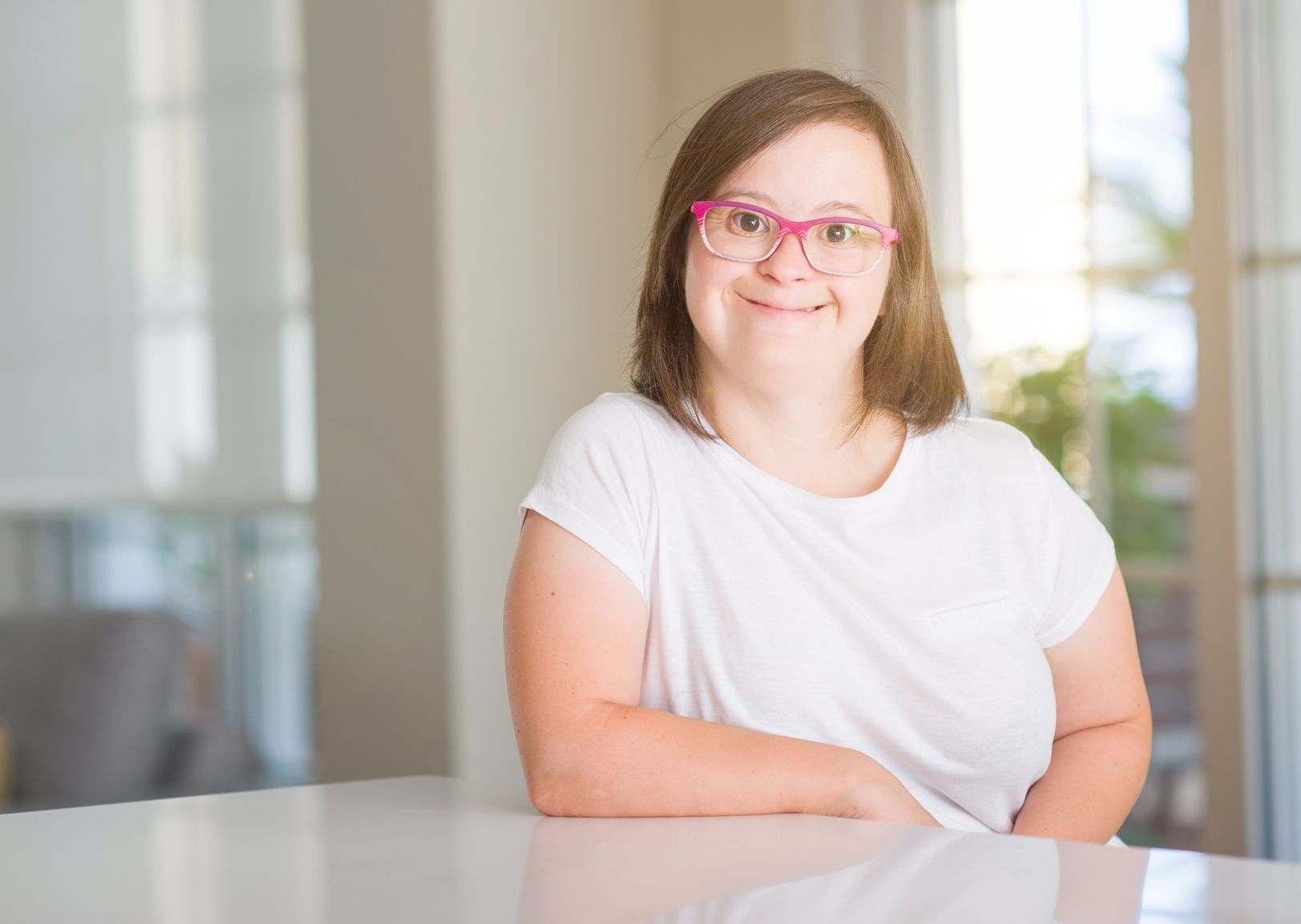 A young girl with down syndrome is sitting at a table wearing glasses and smiling.