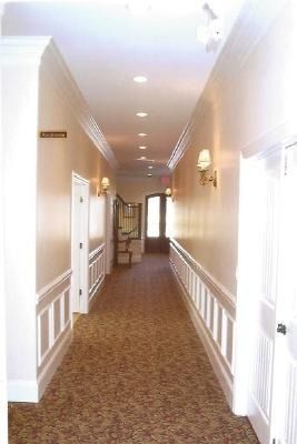 Empty carpeted hallway with white trim and doors, lit by ceiling lights, leading to a window at the far end