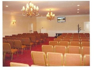 Empty banquet hall with rows of tan chairs, chandeliers, and a small wall-mounted TV.