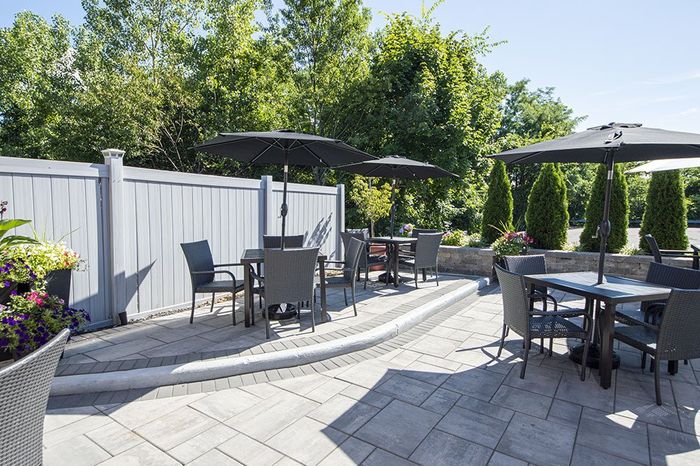 An outdoor patio featuring gray stone pavers, multiple dining tables with dark umbrellas, and chairs near a gray fence.