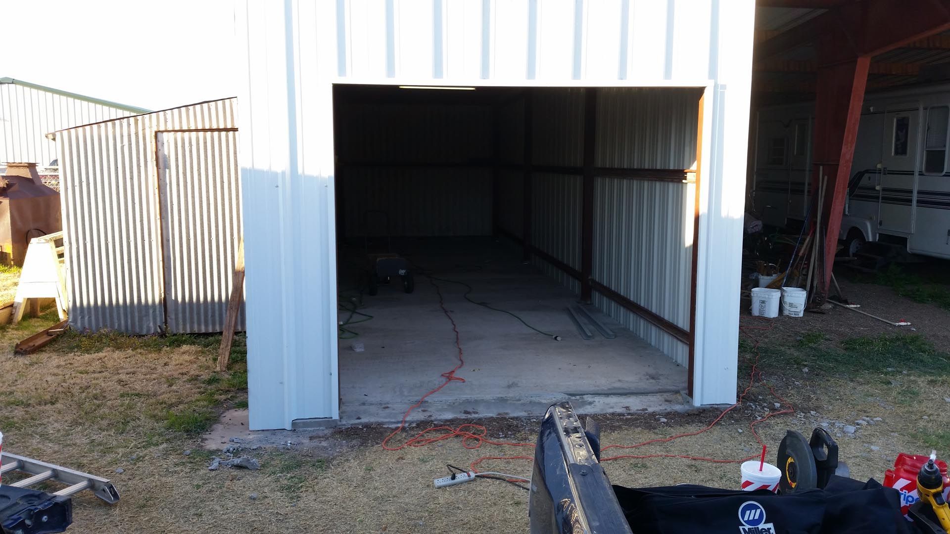 Open metal storage shed with a concrete floor and tools inside, beside a garage in daylight