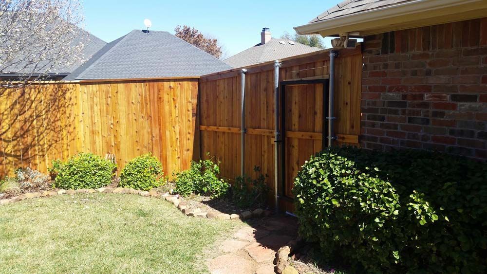 Backyard corner with wooden fence, gate, brick wall, grass, and shrubs in sunlight