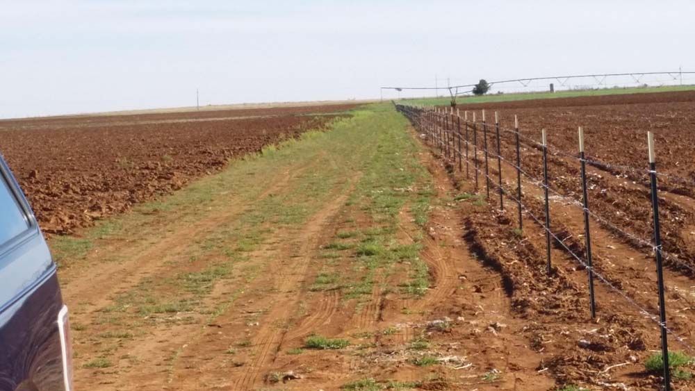 Rural dirt path between plowed fields, bordered by a wire fence under a cloudy sky