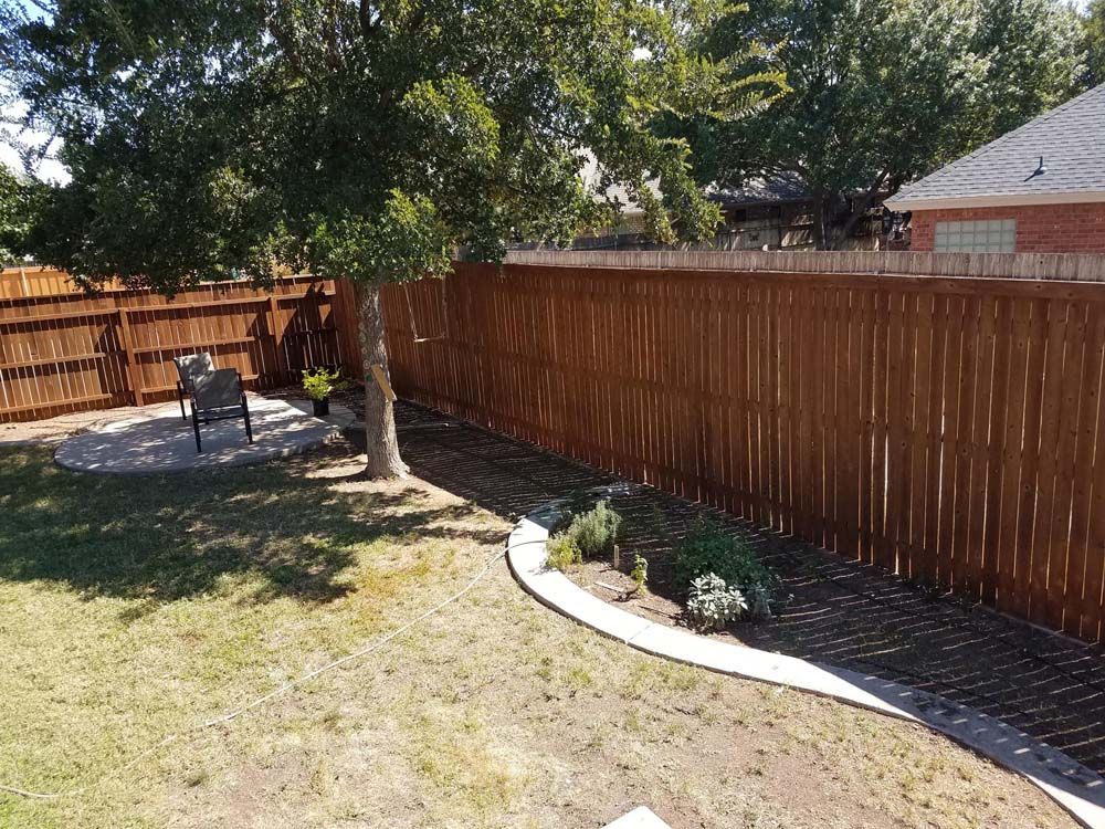Backyard with wooden fence, tree, lawn, and small stone garden bed under bright sun