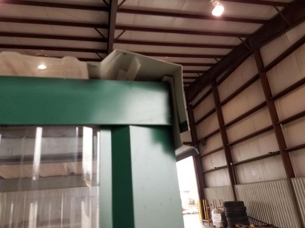 Green industrial machine inside a warehouse with metal roof beams and stacked materials.