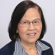 A smiling person with dark hair and glasses wears a blue blazer over a black and white pinstriped collared shirt.