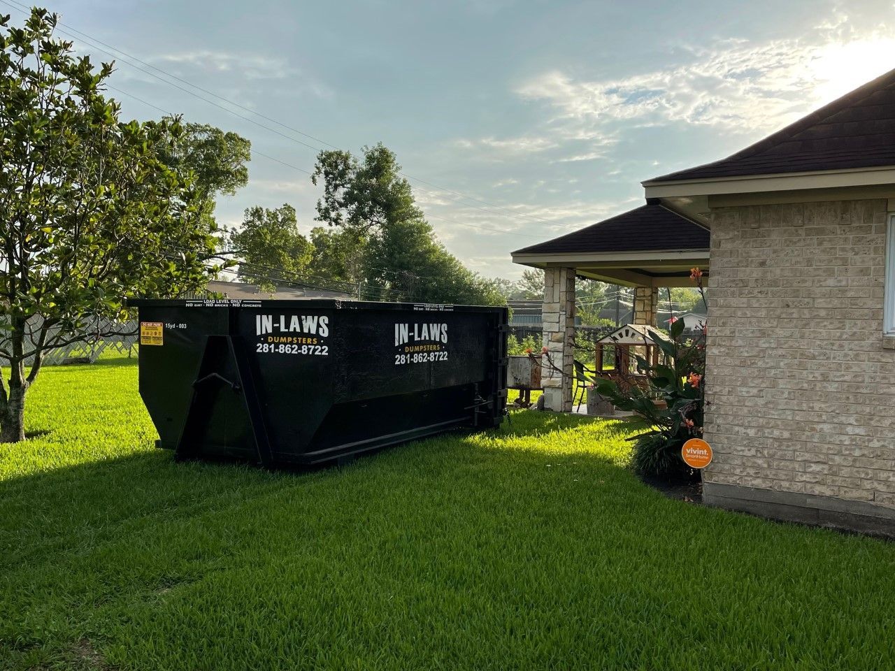 A dumpster is sitting in the grass in front of a brick house.