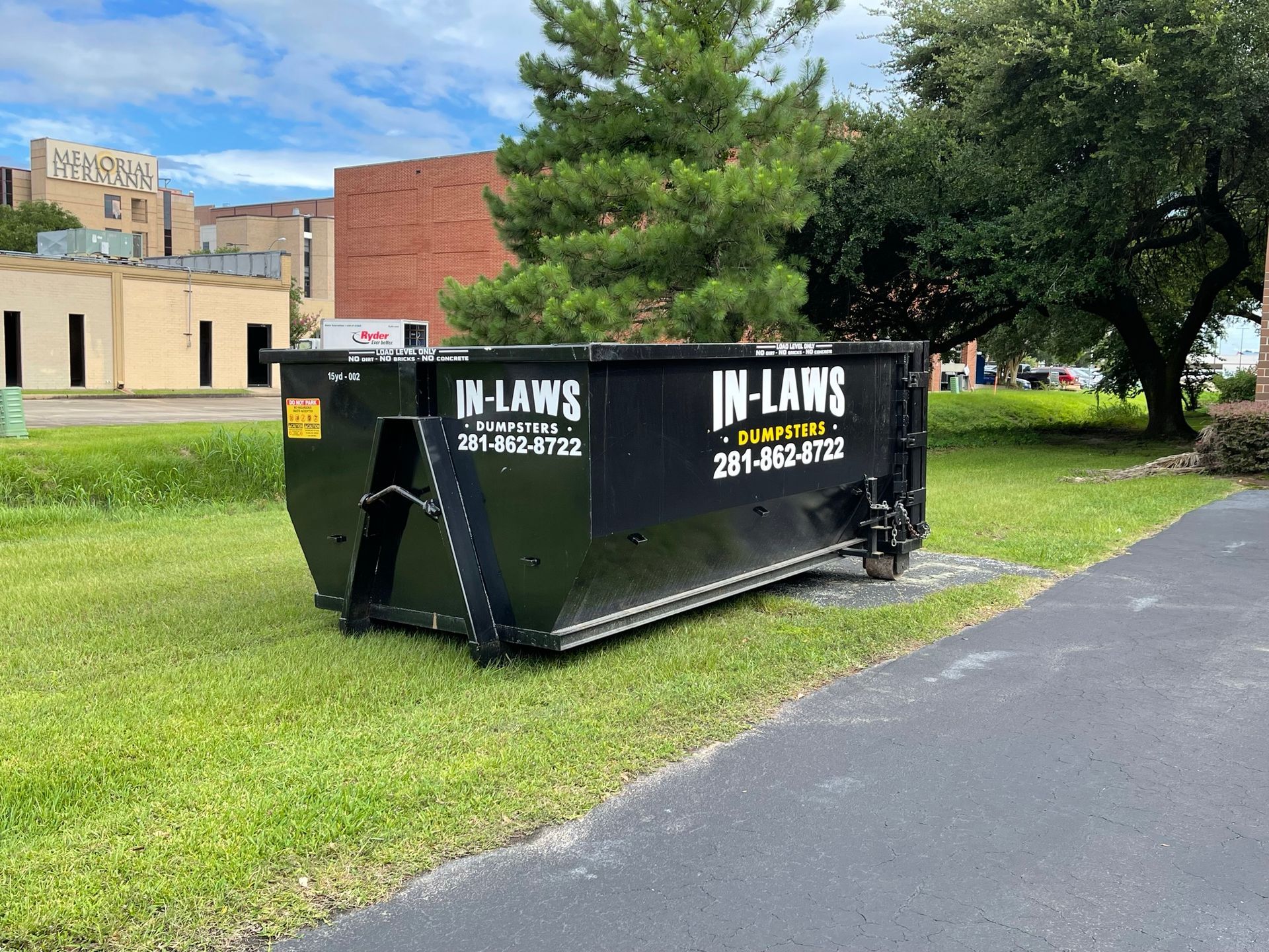 A large black dumpster is parked in the grass next to a road.