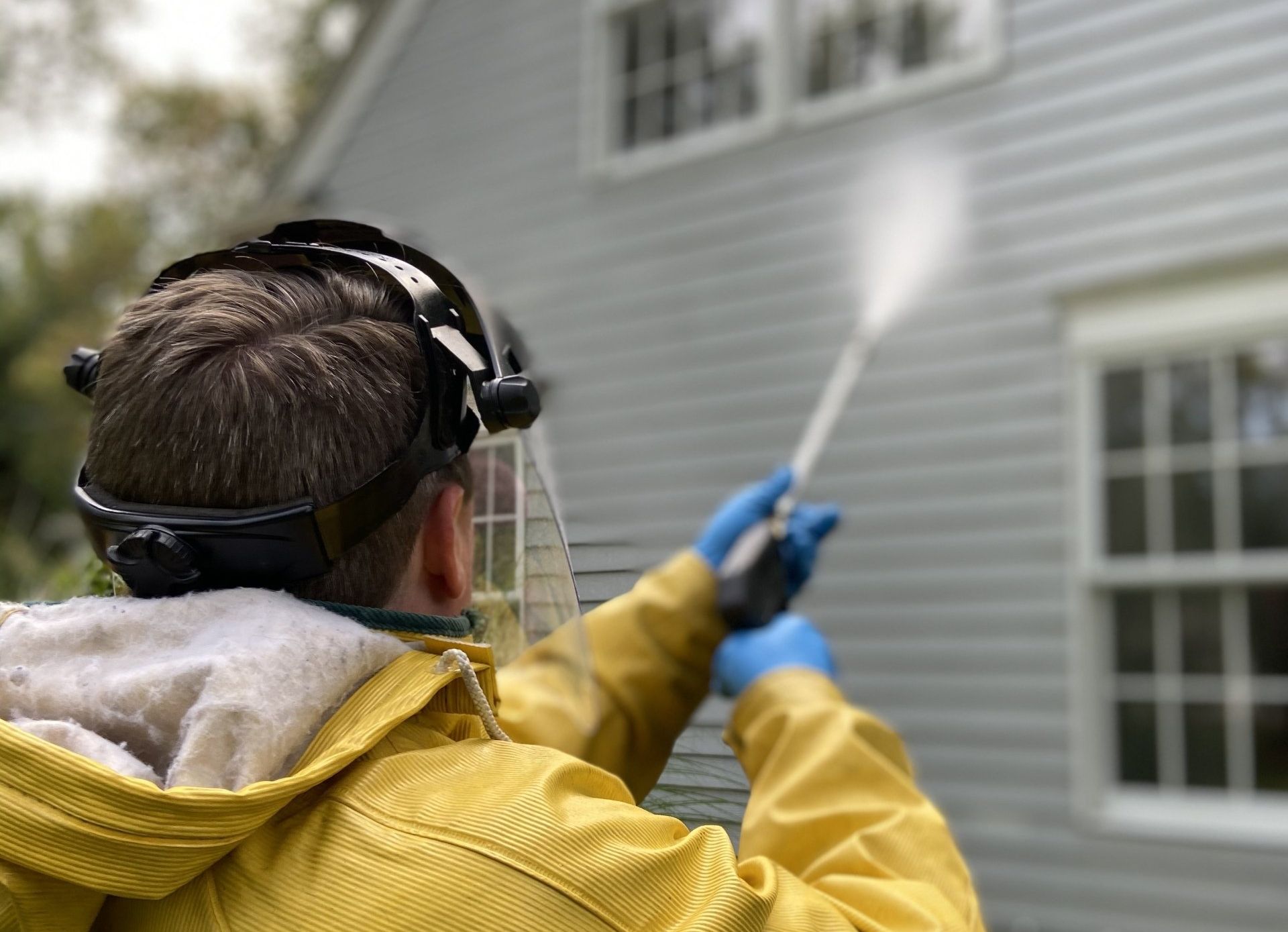 A man in a yellow jacket is cleaning the side of a house with a high pressure washer.
