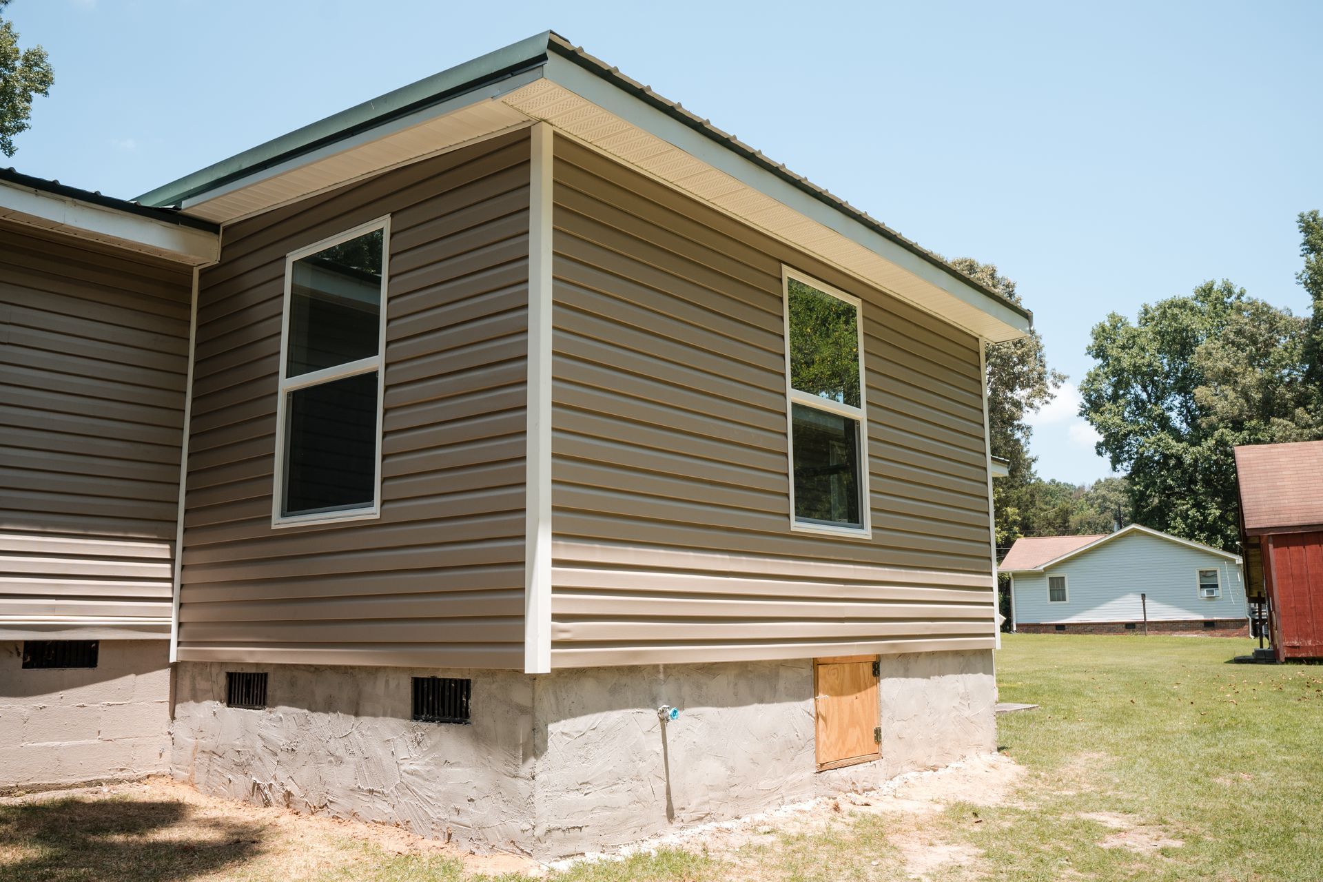 Side view of a tan vinyl-sided house with white trim on a concrete foundation in a grassy yard.