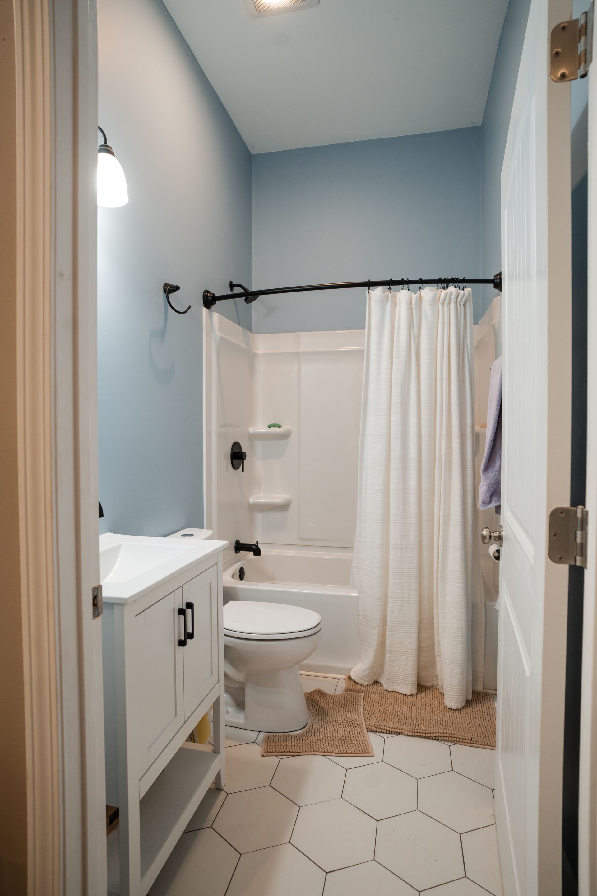 Small bathroom with light blue walls, white vanity, toilet, shower curtain, and hexagon tile floor.