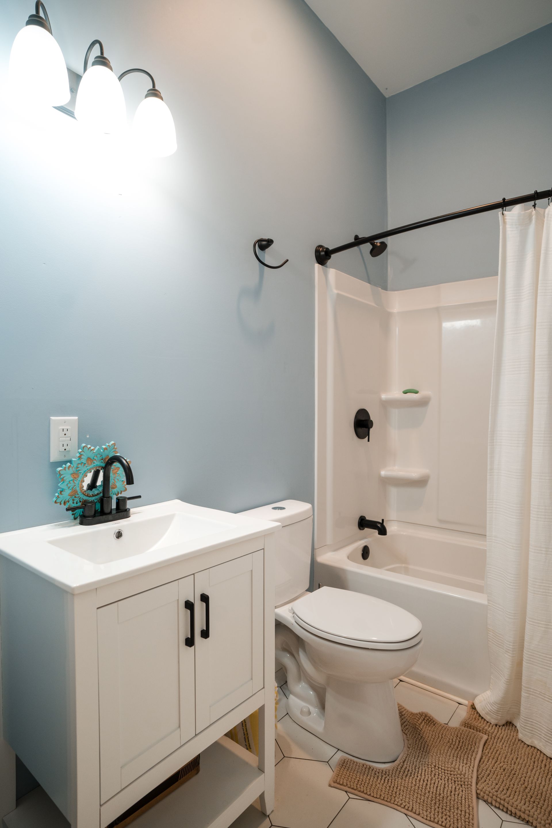 Small bathroom with white vanity, toilet, shower curtain, and woven bath mat against pale blue walls.