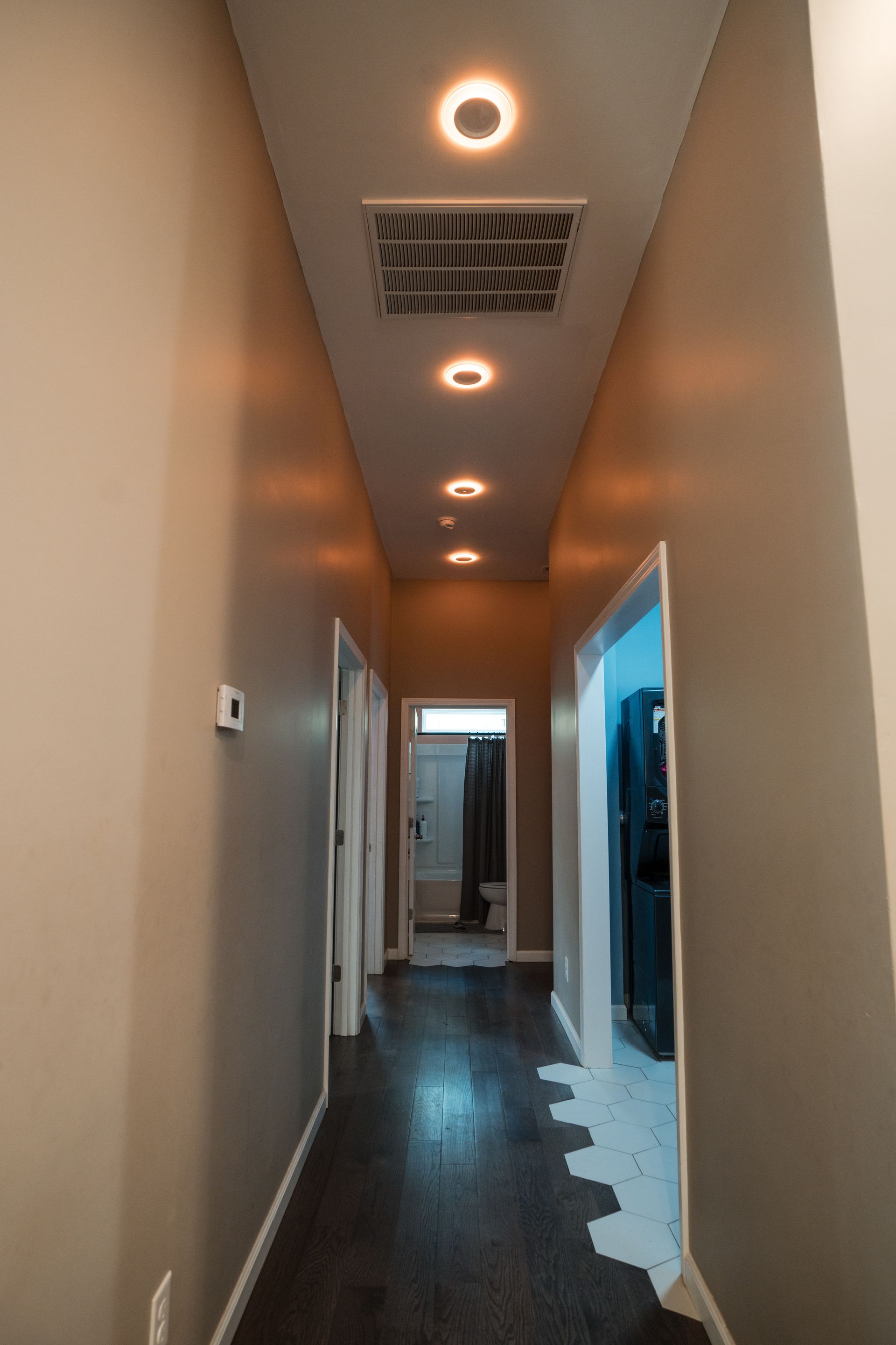 Narrow hallway with dark wood floor, beige walls, recessed ceiling lights, and a doorway at the end.