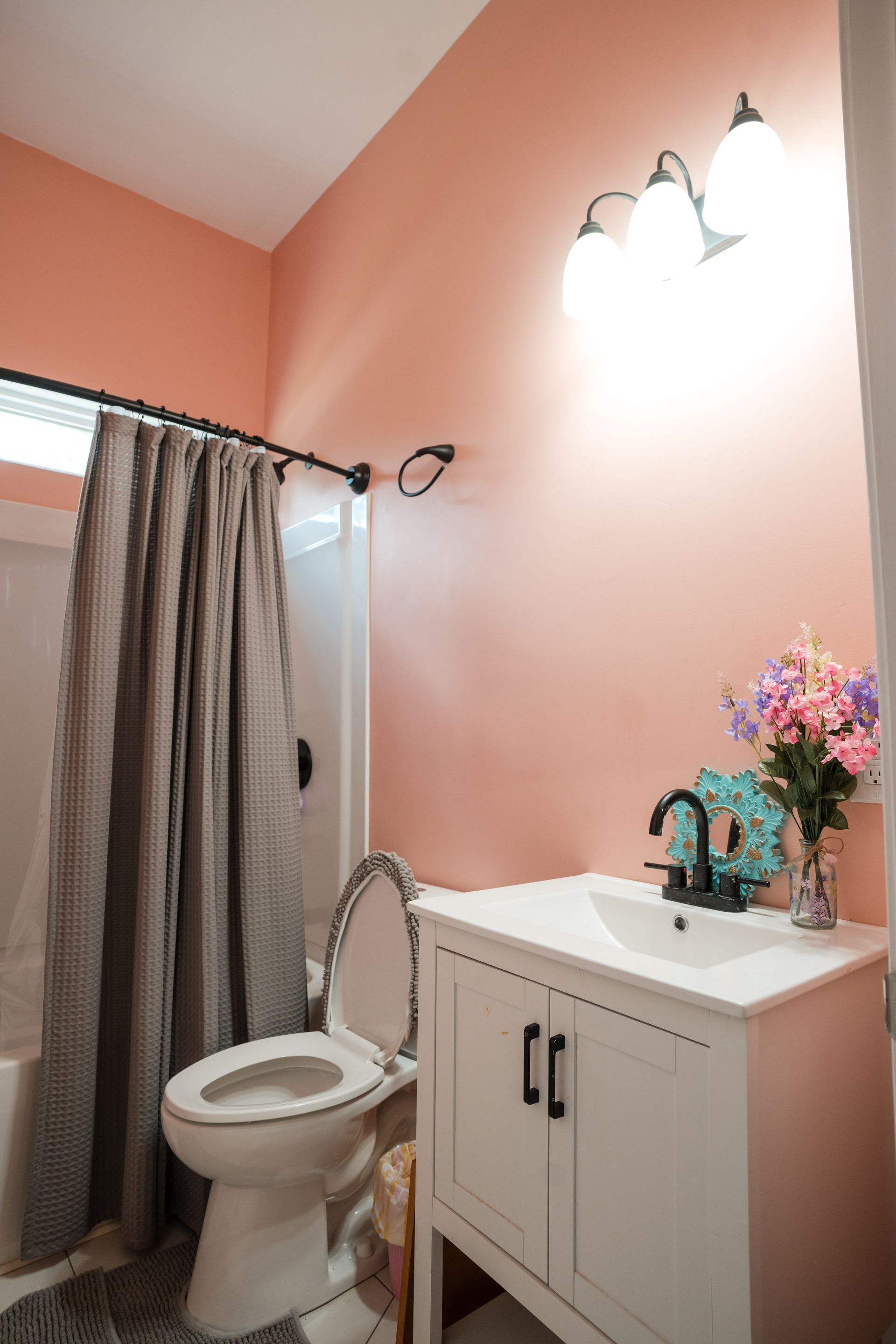 Small bathroom with peach walls, white sink cabinet, toilet, shower curtain, and floral decor.