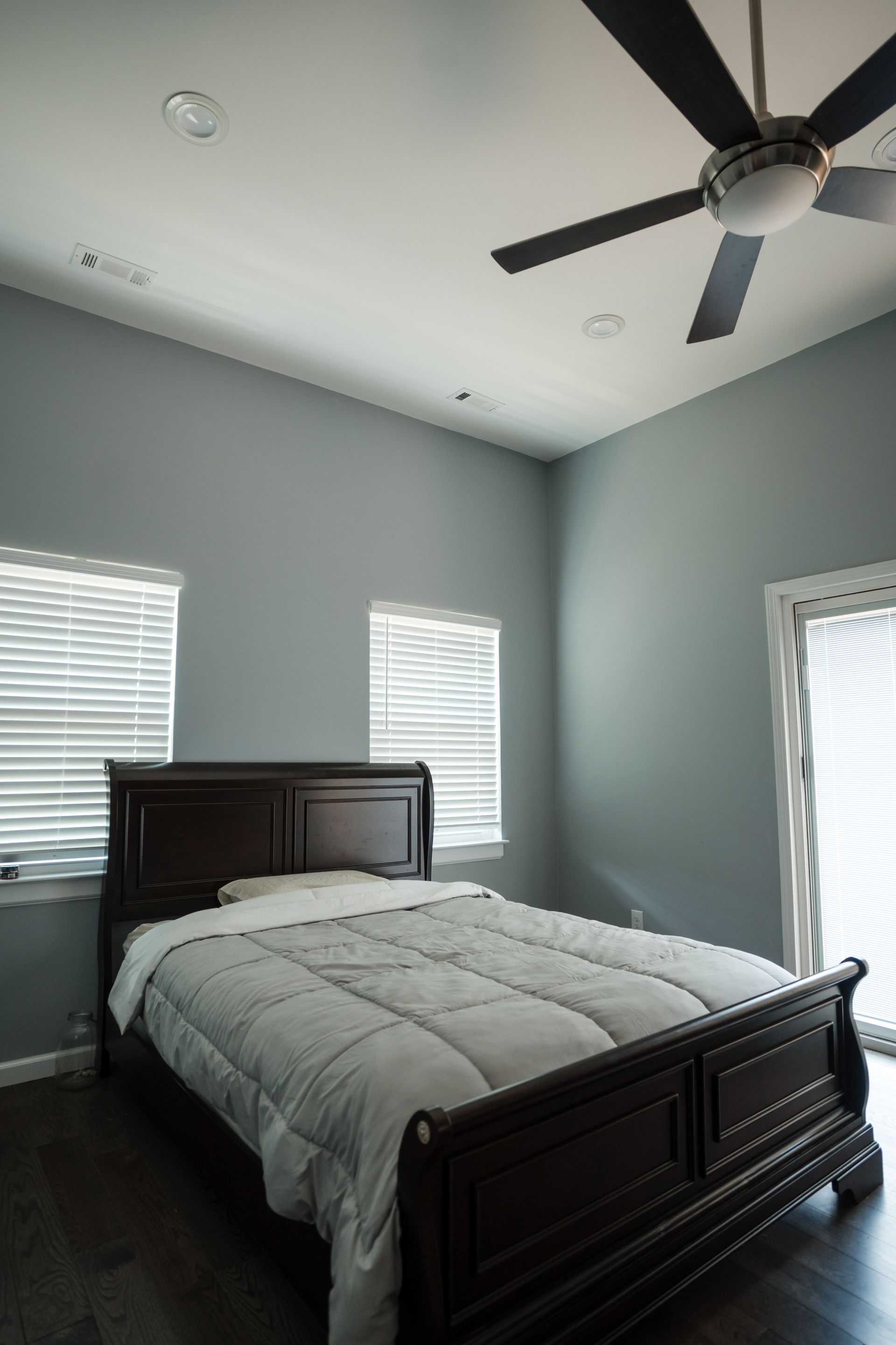 Bright bedroom with dark wood bed, gray walls, white bedding, windows, and a ceiling fan