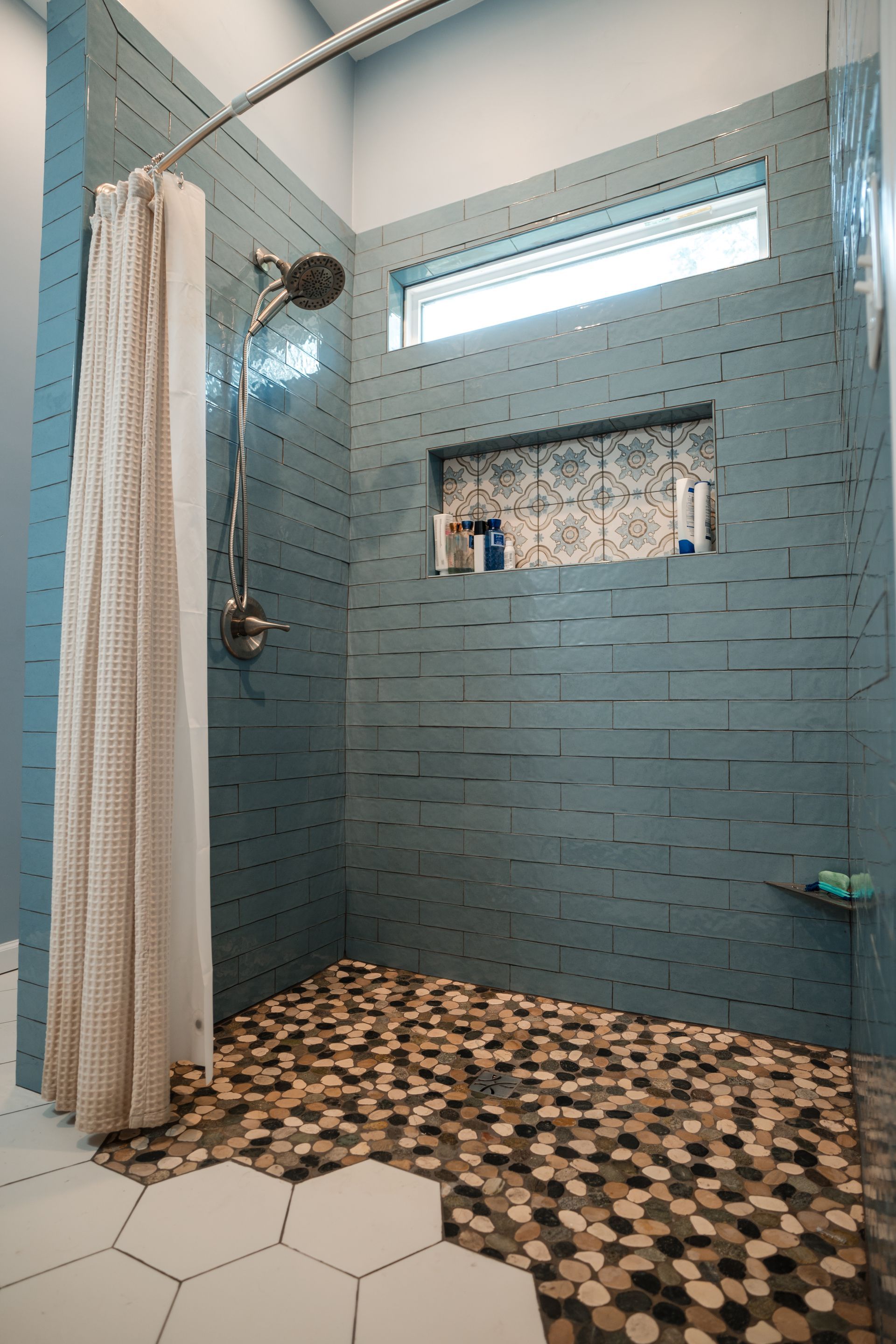Blue-tiled shower with pebble floor, glass partition, and white curtain.