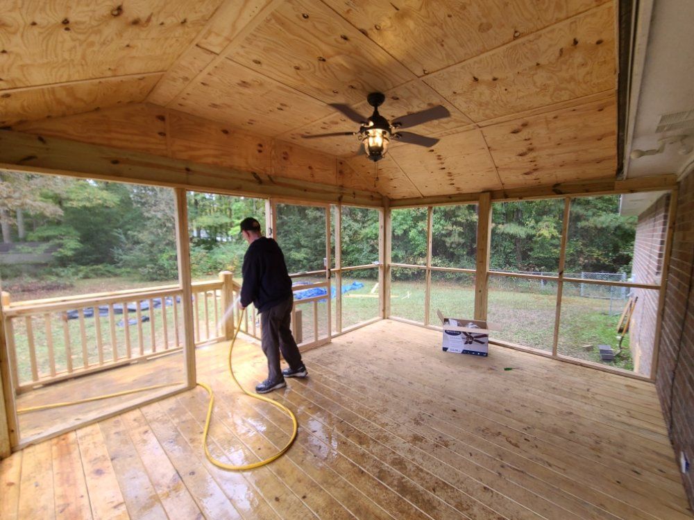 Person sanding a wooden screened porch floor with a yellow hose nearby and forest view outside.
