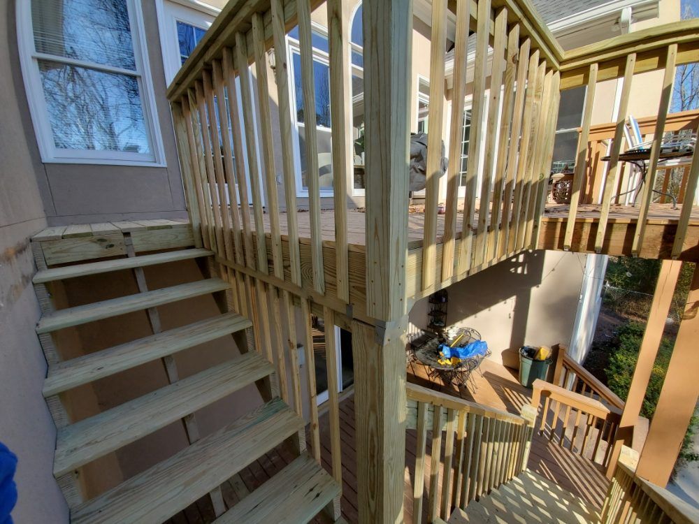 Wooden staircase and deck railings beside a house, viewed from an upper landing looking down to a lower patio.