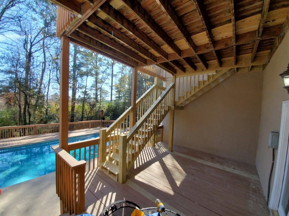 Covered backyard deck beside a turquoise pool, with wooden stairs and railing in bright sunlight.